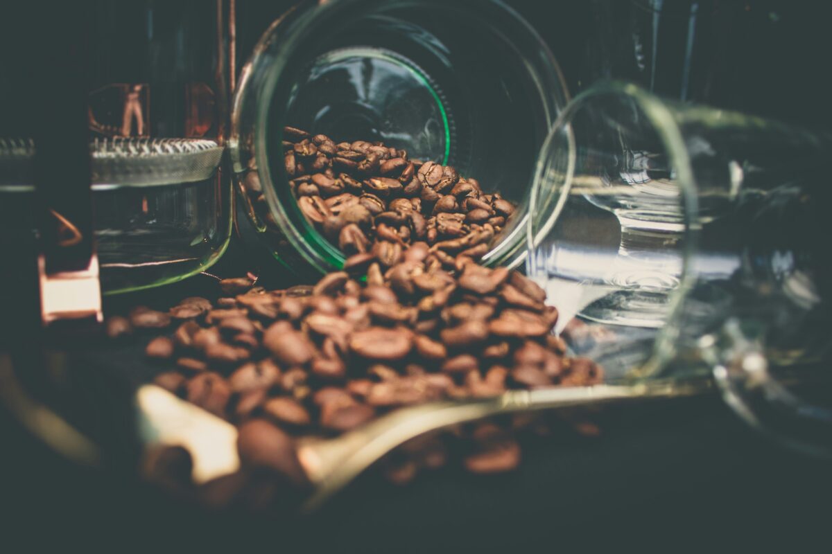 Close-up view of decaf coffee beans spilling out of a glass jar, showcasing their rich, dark brown color and texture, with additional jars and a scoop in the background.